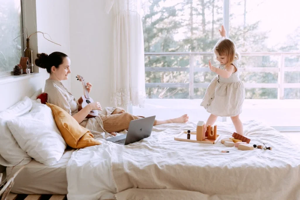 Mother plays ukulele in bed while young daughter jumps around